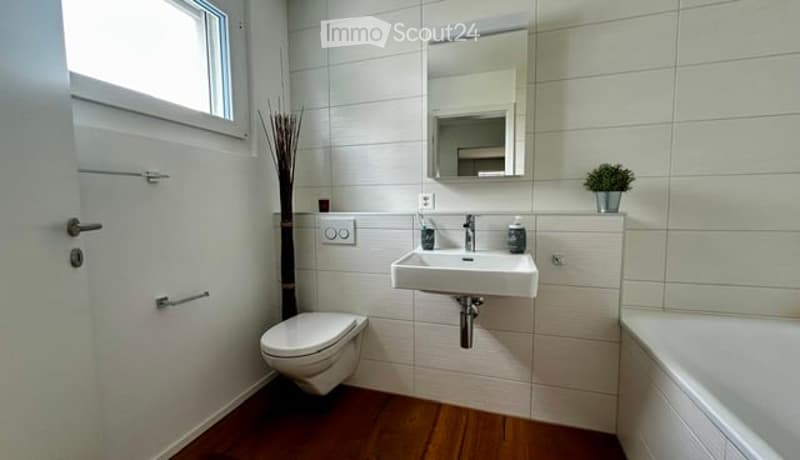 Modern bathroom with white tiles, wooden floor, toilet, sink, mirror, and bathtub.