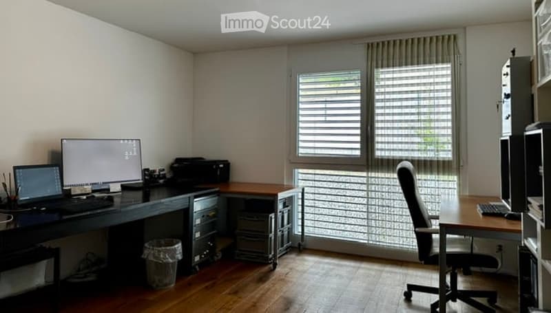 A modern, well-lit home office with wooden flooring, large windows with blinds, and a desk setup featuring a laptop, printer, and office chair.