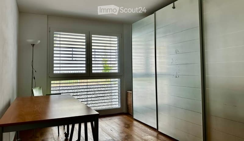 Bright, minimalist room with wooden flooring, large window with blinds, a simple wooden table and chair, and a sleek white sliding door.