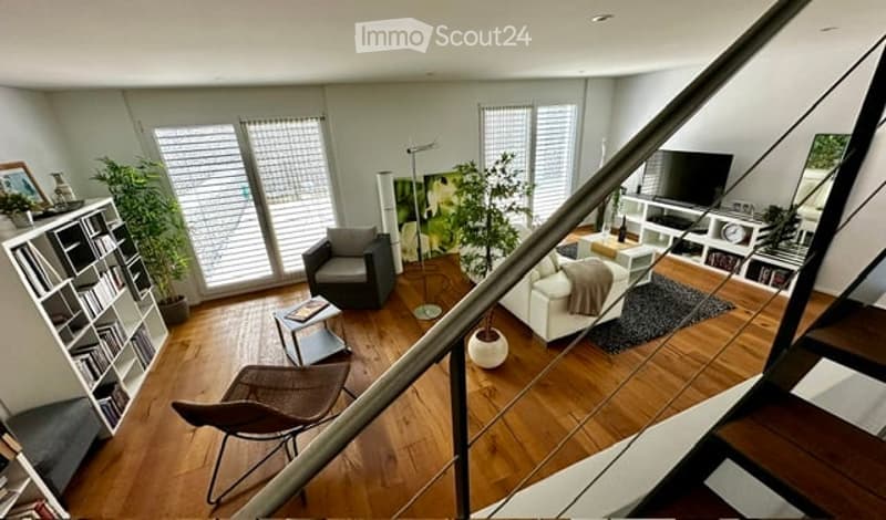Modern living room with hardwood floors, large windows, white sofa, grey armchair, coffee table, bookshelf, floor lamp, and TV stand.