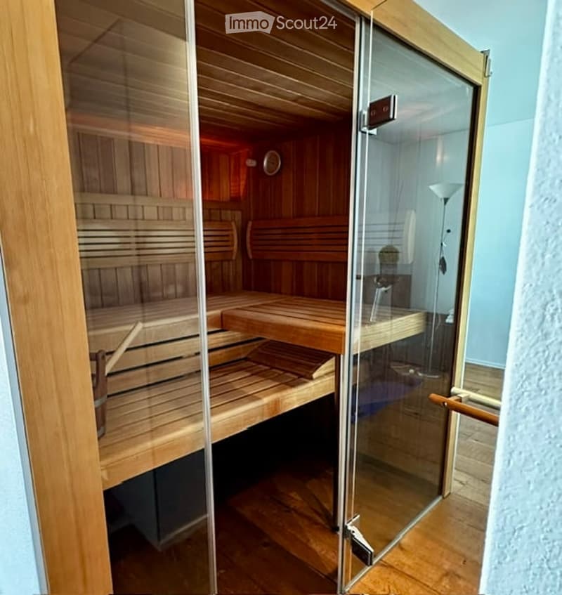 A wooden sauna with multiple benches, glass door, and a floor lamp visible in the background.