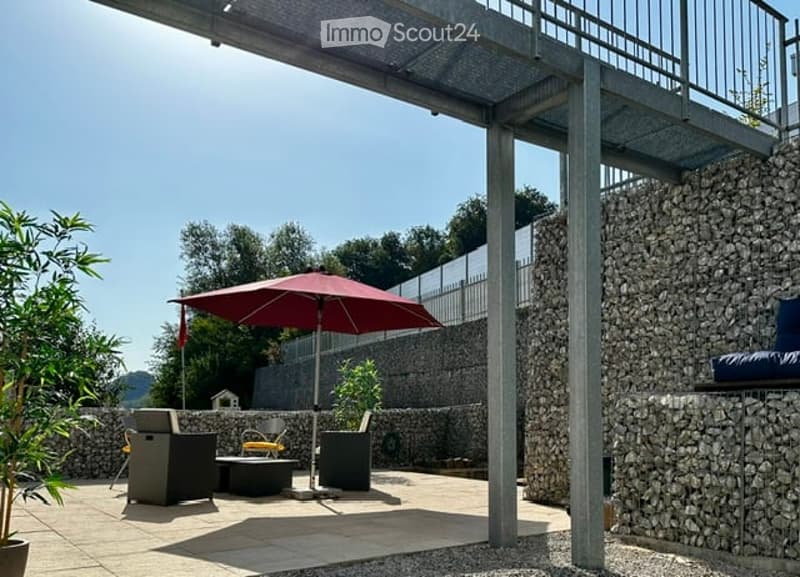 Outdoor patio with a large umbrella, seating area, and modern furniture, surrounded by stone walls and greenery.