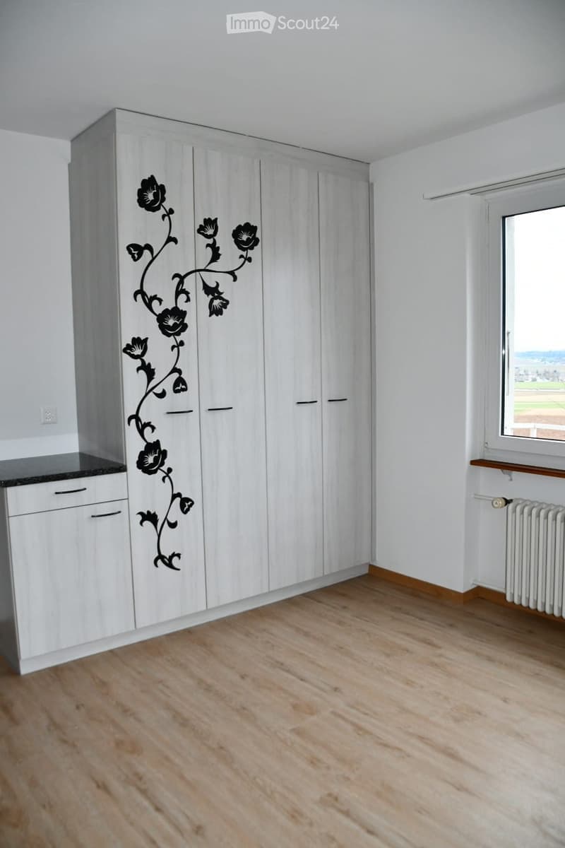 Empty room with light wooden flooring, white walls, large built-in wardrobe with floral design, window with view, radiator, and small countertop with drawers.