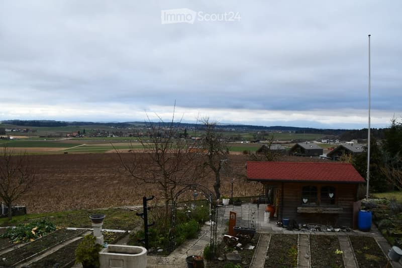 Garden area with a small wooden shed, a flagpole, and a view of a rural landscape with fields and distant hills.