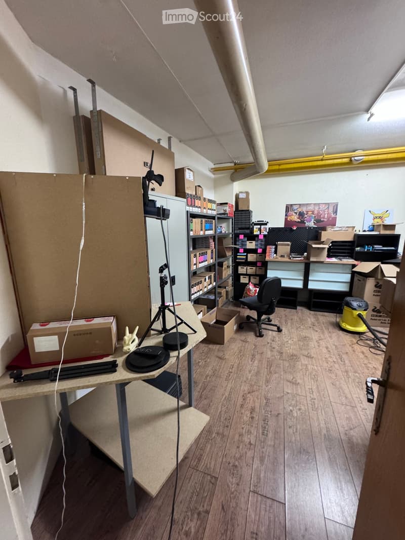 A room with wooden flooring, a table with various items, a chair, shelves filled with boxes and items, a tripod, and a vacuum cleaner. The room appears to be used for storage and work.