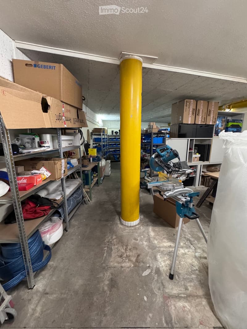 Empty storage room with concrete floor, metal shelving, and a central yellow pillar. Various boxes and tools are stored on shelves and the floor.