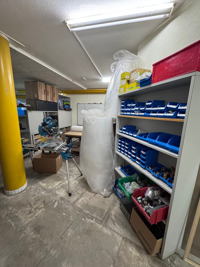 Workshop or storage area with concrete floor, shelves filled with tools and supplies, a saw on a stand, and various boxes and containers.