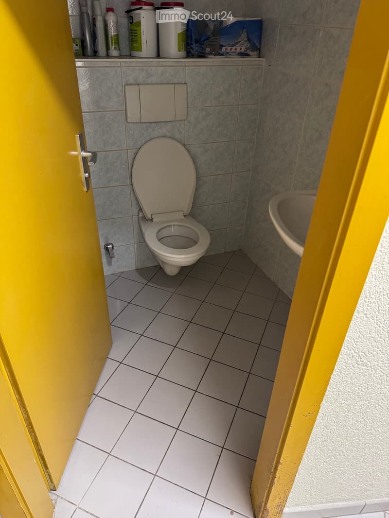Small bathroom with a toilet, sink, and toilet paper holder. Grey tiled walls and white tiled floor.