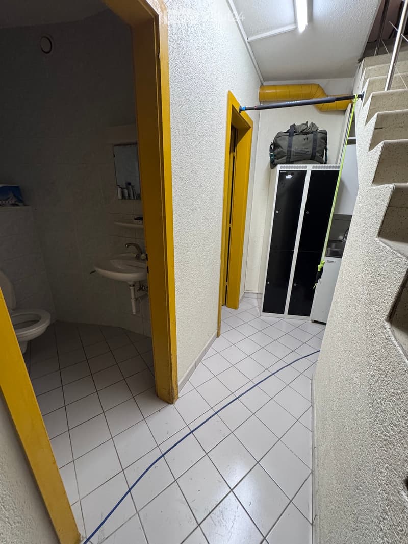 A narrow hallway with white tiled flooring, leading to a small bathroom and a storage area. The bathroom contains a sink, toilet, and a mirror. A black storage cabinet is visible in the hallway, with a duffel bag placed on top.