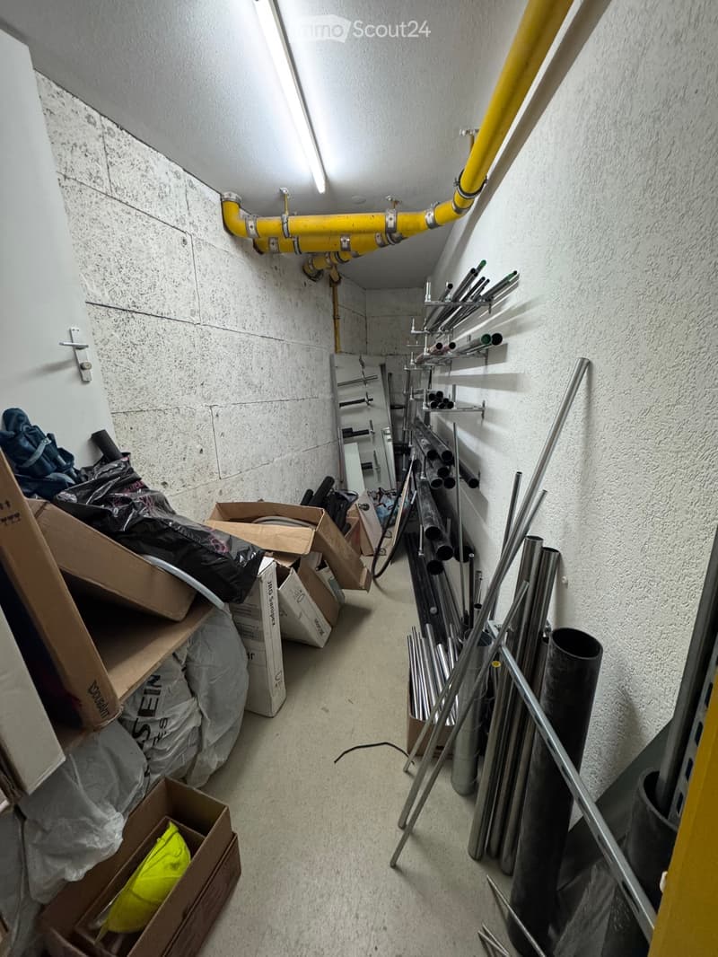 A narrow, unfinished corridor with exposed pipes, storage boxes, and metal rods. The walls are concrete, and the floor is concrete. There are shelves with pipes and tools, and a ladder is visible in the background.