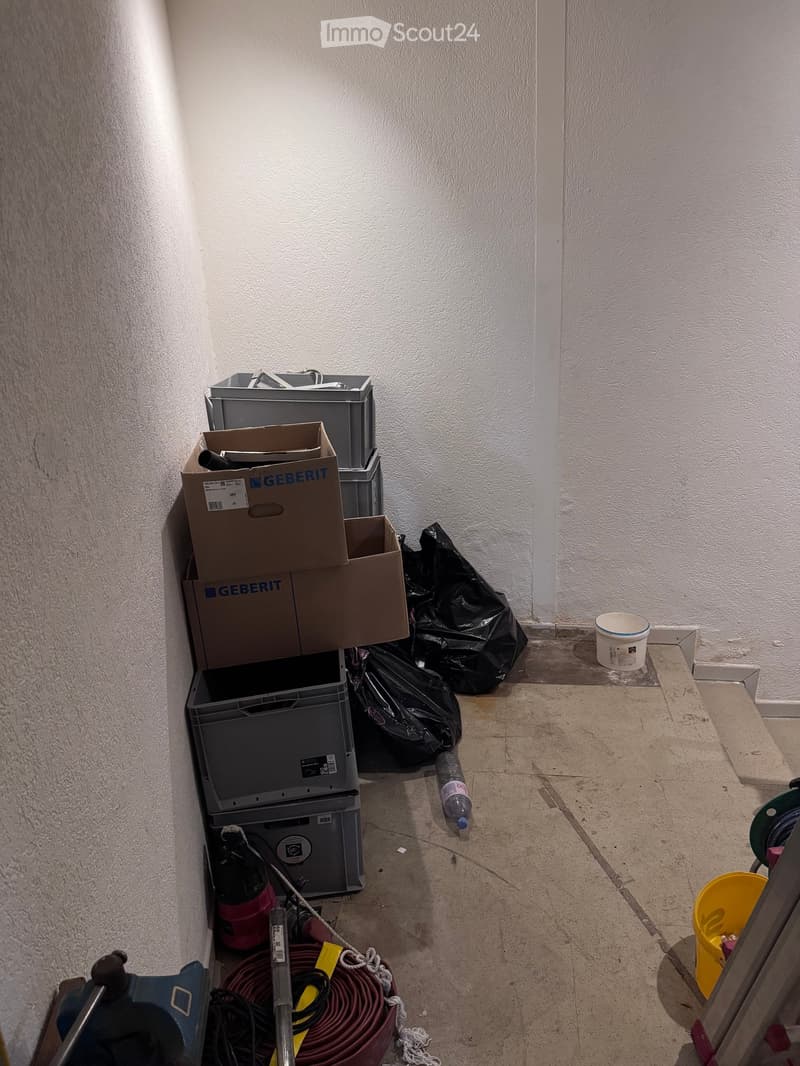 Empty room with bare walls and tiled floor, containing scattered items like boxes, a water bottle, and a bucket.