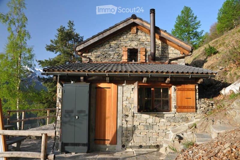 A rustic stone cottage with a wooden door, small windows, and a chimney, situated in a mountainous area with a wooden railing and steps leading up to it.