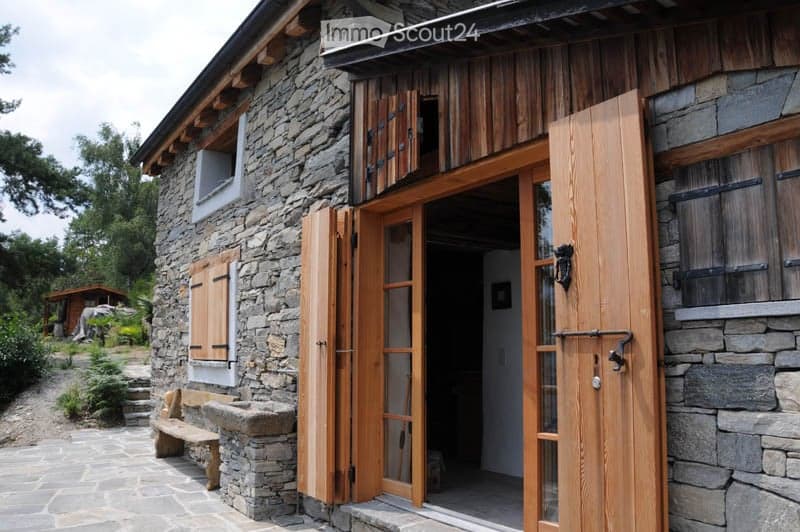 A rustic stone house with wooden shutters and a large wooden door, featuring a stone bench and a small garden area.