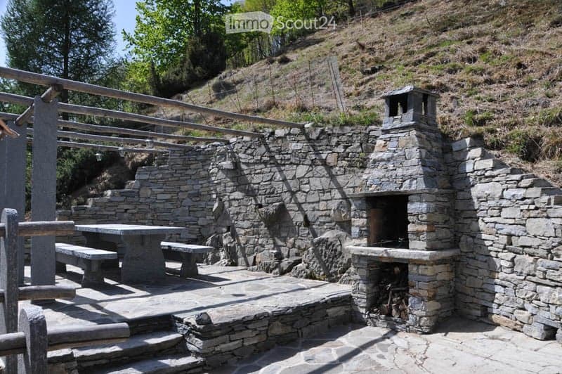 Stone outdoor seating area with a built-in stone fireplace, a wooden railing, a stone table, and benches, surrounded by greenery and a hillside.