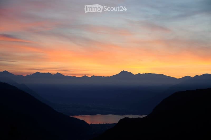 Panoramic view of a mountain range at sunset, with a lake in the foreground and a cityscape visible in the distance.