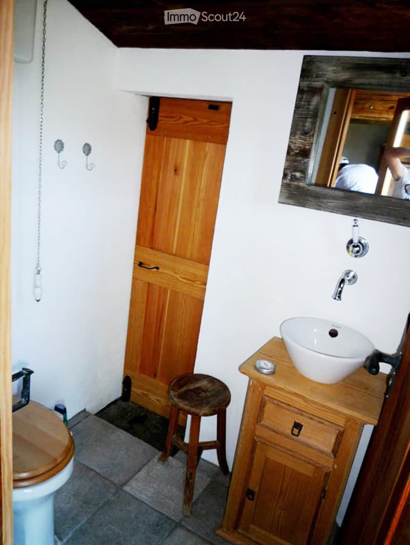 Cozy bathroom with a wooden door, a toilet, a sink on a wooden cabinet, a small wooden stool, and a mirror above the sink.