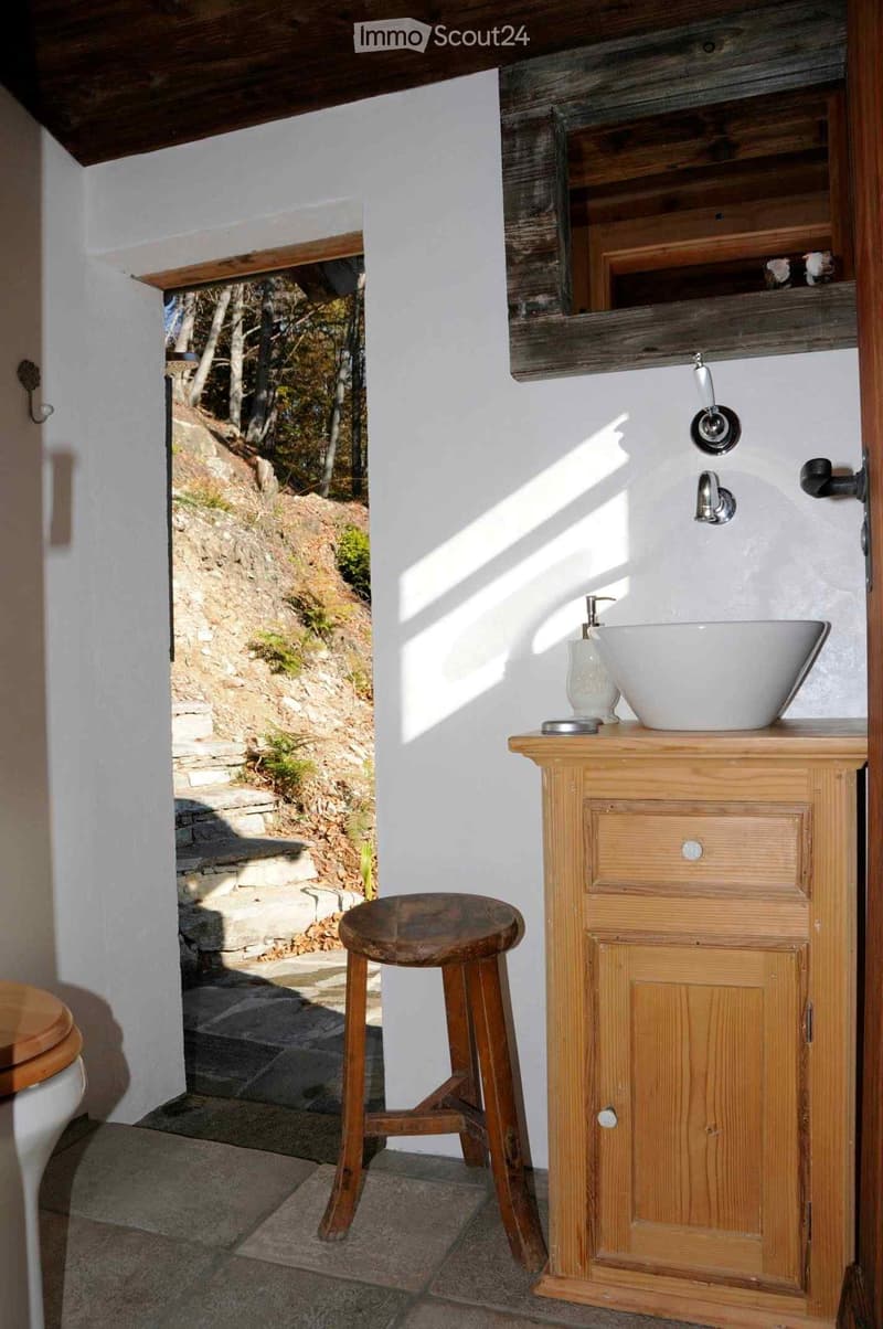 Bathroom with a wooden vanity, sink, faucet, and a wooden stool. A toilet is visible to the left, and the room has a view of a stone pathway leading outdoors.