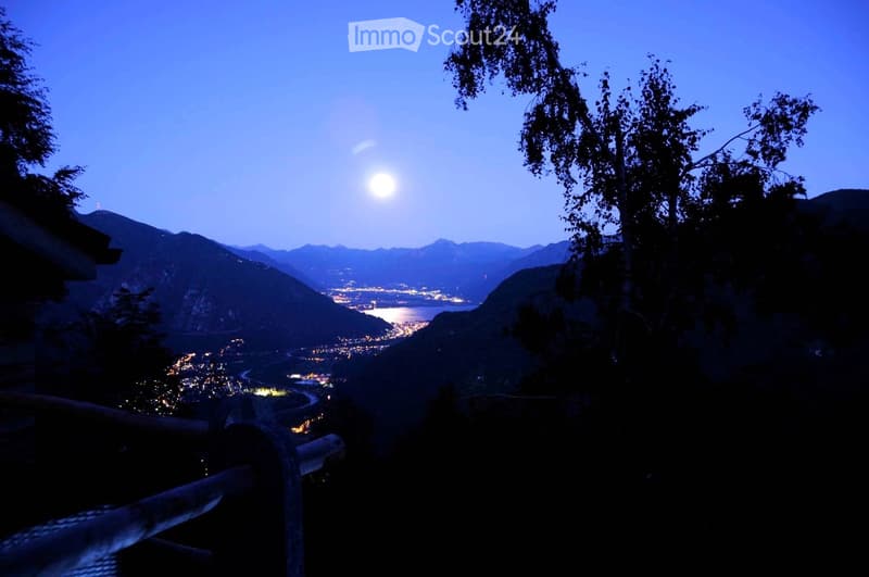 Nighttime view from a balcony or terrace overlooking a town with lights, surrounded by mountains and a body of water.