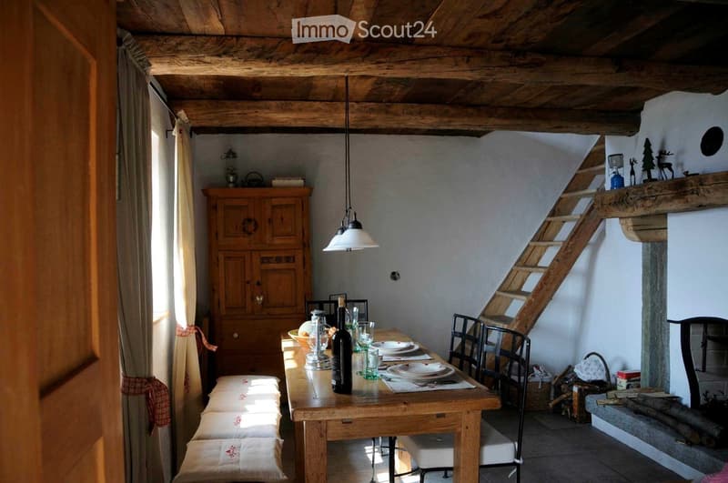 Cozy kitchen with wooden dining table, chairs, and a wooden cupboard. The room features exposed wooden beams, a staircase, and a window with a curtain.