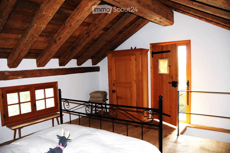 Cozy attic bedroom with wooden beams, slanted ceiling, large window, bed with white linens, wooden wardrobe, bench, and a small stool.