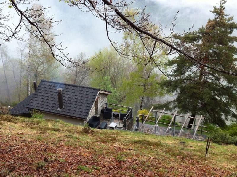 A small, single-story house with a dark tiled roof and a chimney, situated on a grassy slope surrounded by trees in a misty forest setting.