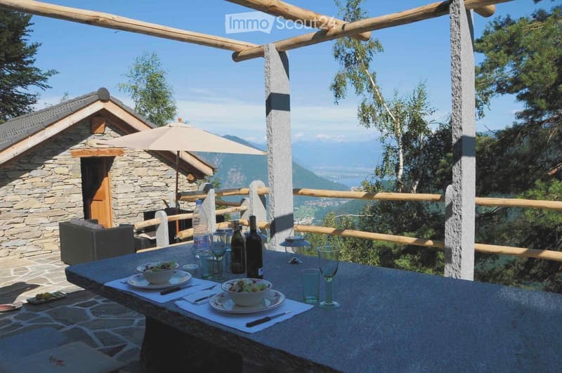 Outdoor terrace with a stone table set for dining, overlooking a scenic mountain view with a stone house in the background.