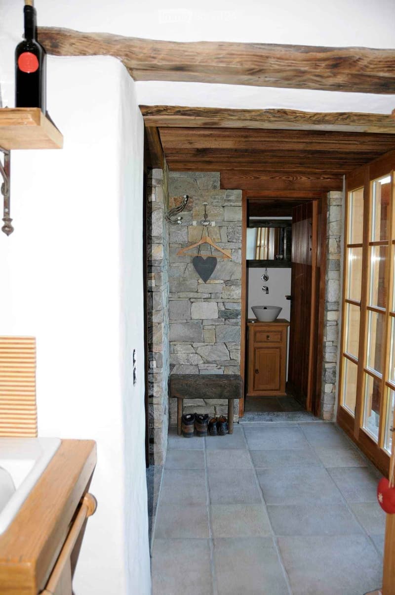 A rustic hallway with exposed wooden beams, stone walls, and a wooden bench. Visible items include a coat hanger, shoes, a sink, and a wooden cabinet.