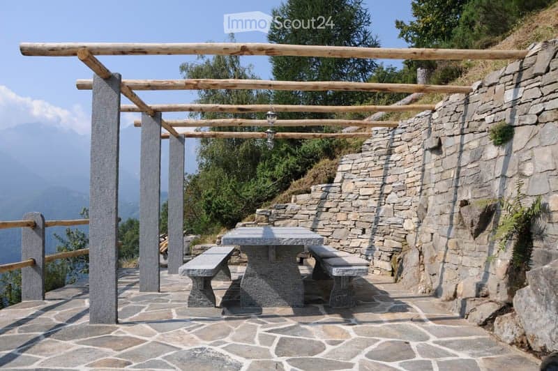 Outdoor seating area with stone table and benches under a wooden pergola, overlooking a scenic mountain view.