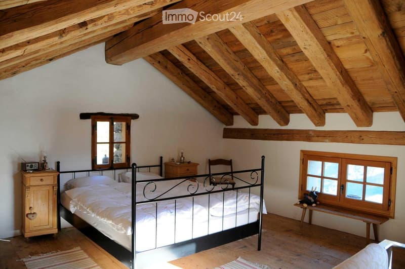 Cozy attic bedroom with wooden beams, a metal-framed bed, nightstand, chair, bench, and a small table with a toy on it.