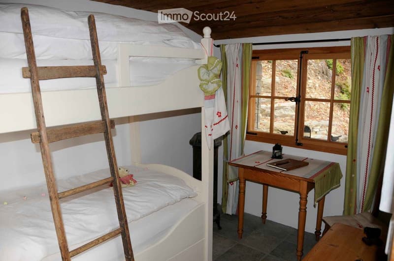 Cozy bedroom with a wooden bunk bed, a small wooden table with a lamp, a chair, and a stool. The room features wooden beams on the ceiling and a window with curtains.