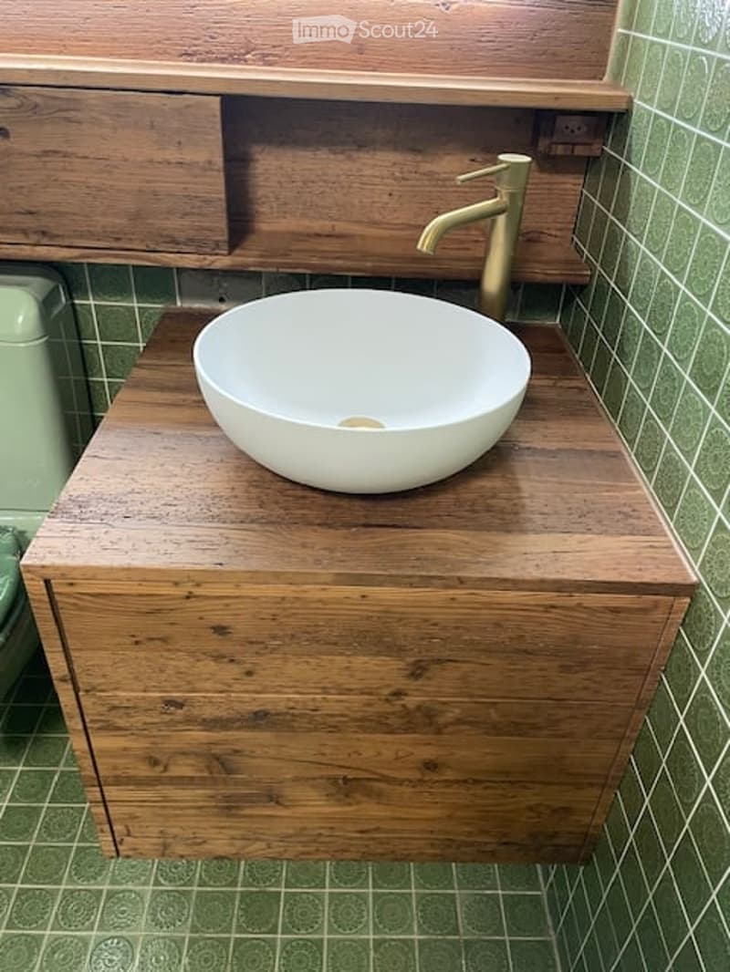 A wooden bathroom vanity with a round white sink and a brass faucet, mounted on a tiled wall with a green pattern.