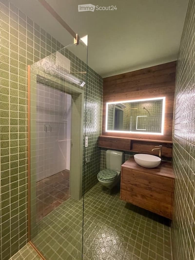 green tiles, shower, white toilet, white sink, wooden counter, mirror, led light on mirror