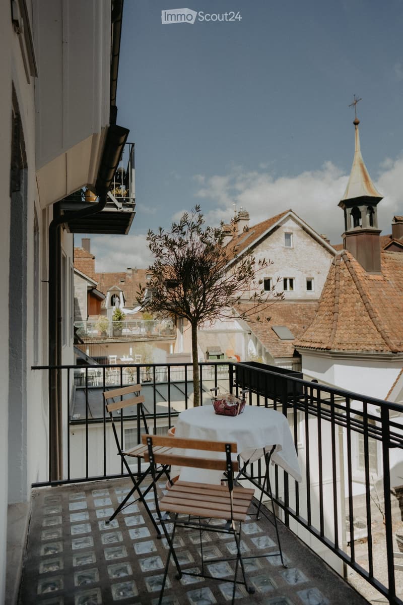 small balcony with black railings, 2 foldable chairs, table with white cloth, potted plant, city view