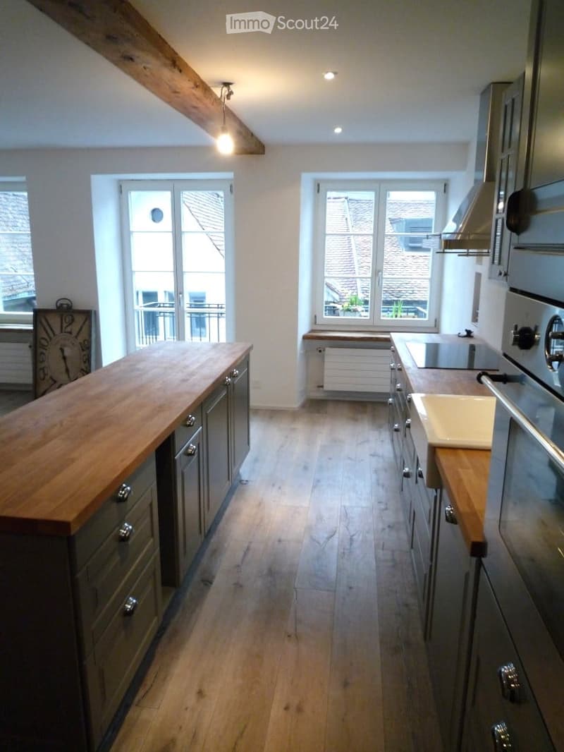 Kitchen with wooden countertop, hanging ceiling lamp, grey cabinets, large windows, wooden floor, and clock on the wall