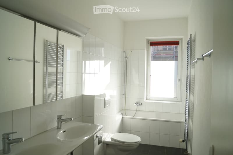 Modern bathroom with white tiles, a large mirror, a sink with a chrome faucet, a toilet, a bathtub, and a radiator. A window with a red blind allows natural light to enter.