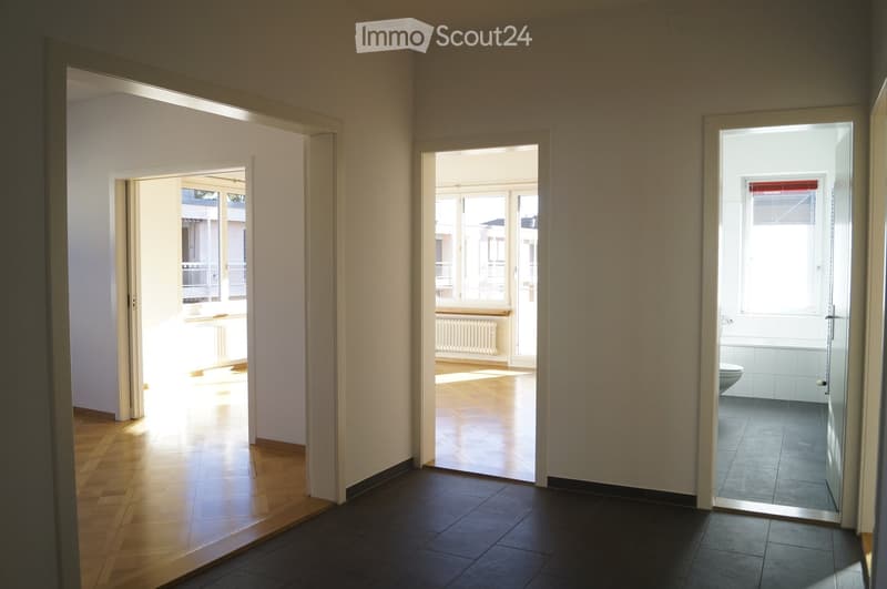 Empty hallway with wooden flooring, leading to a bathroom and another room with large windows and a view of the outside.