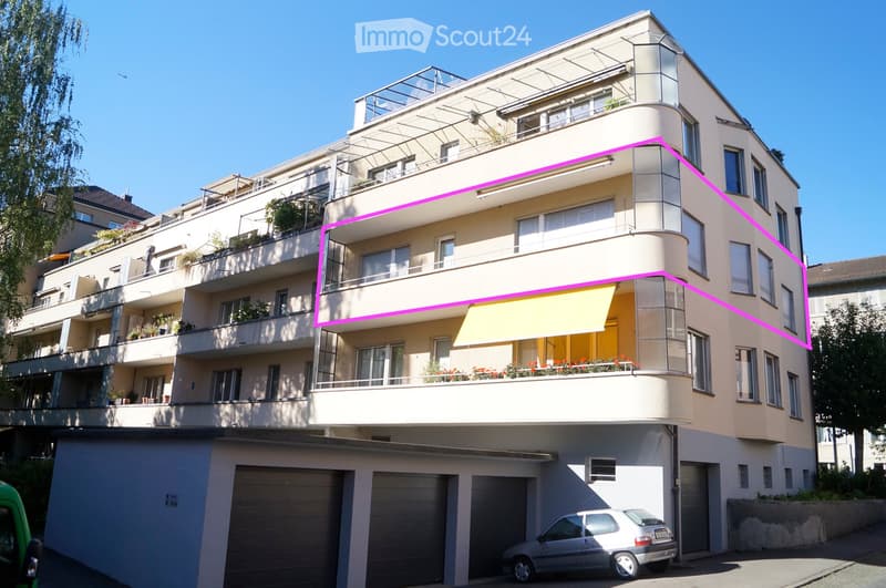 Modern multi-story residential building with balconies, large windows, and a covered parking area. Features include a yellow awning on one balcony and potted plants on multiple levels.