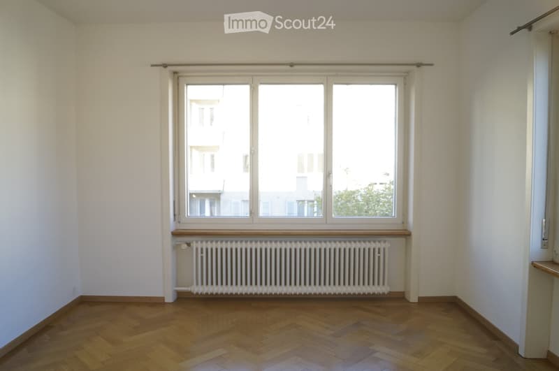 Empty room with wooden flooring, white walls, large window with wooden sill, and radiator below the window.
