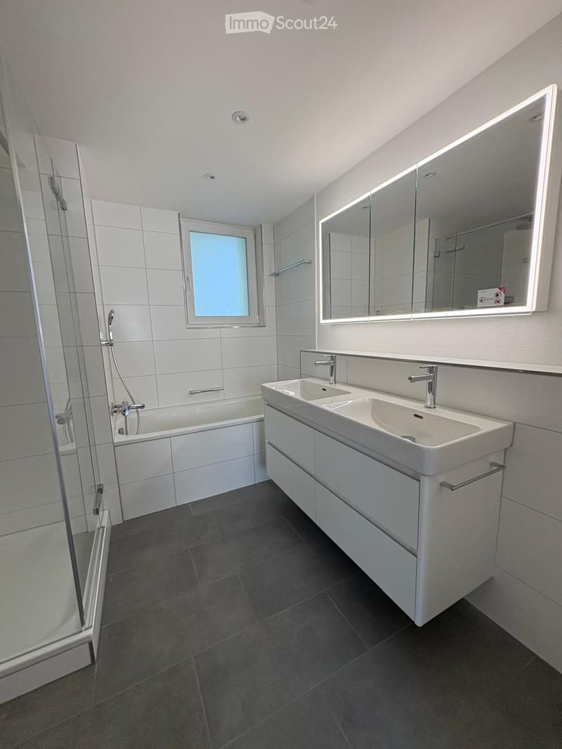 Modern bathroom with a bathtub, double sink, large mirror, and a window providing natural light.
