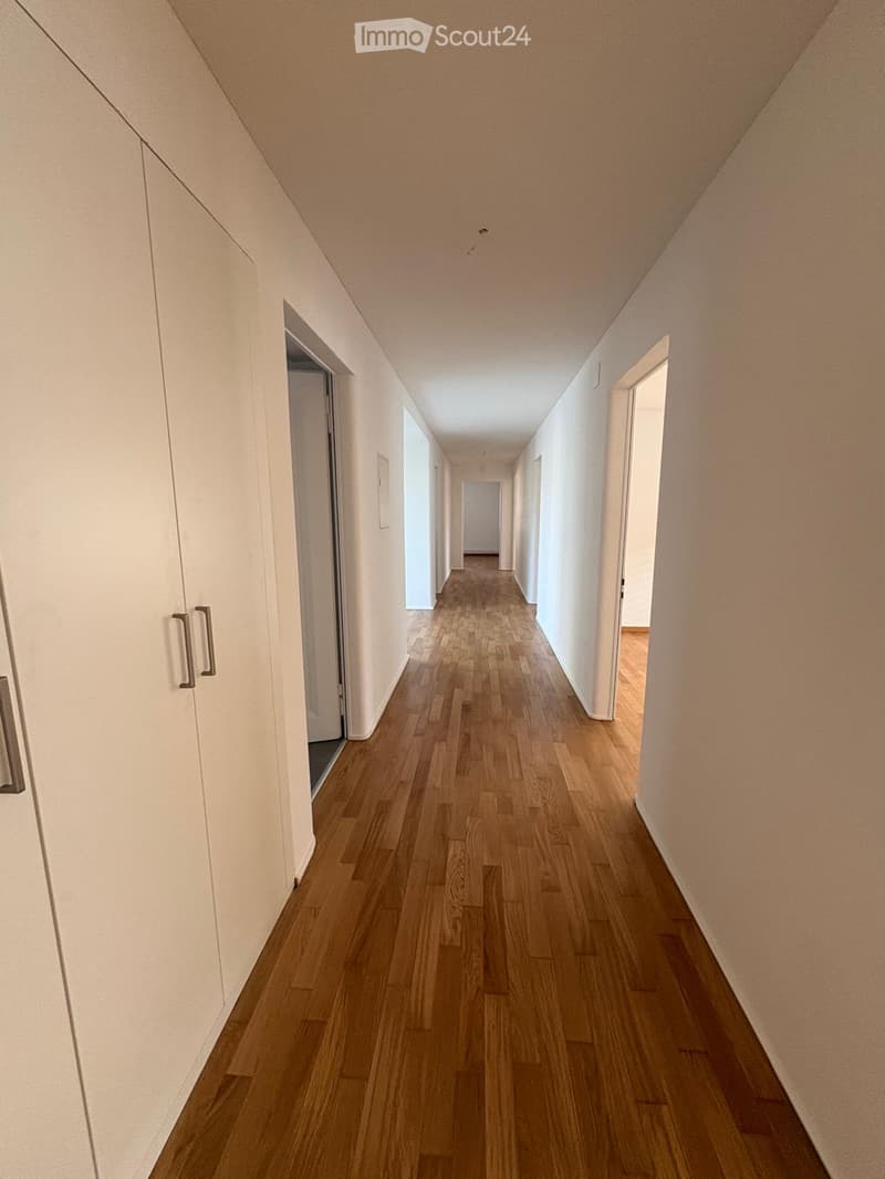 A long, narrow hallway with wooden flooring and white walls, featuring several doors and a slanted ceiling.