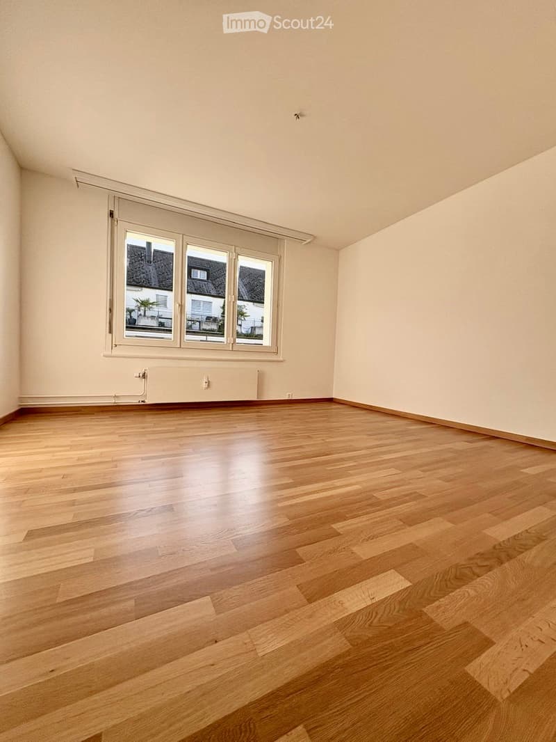 Empty room with wooden flooring, large window with white frame, radiator under the window, and light-colored walls.