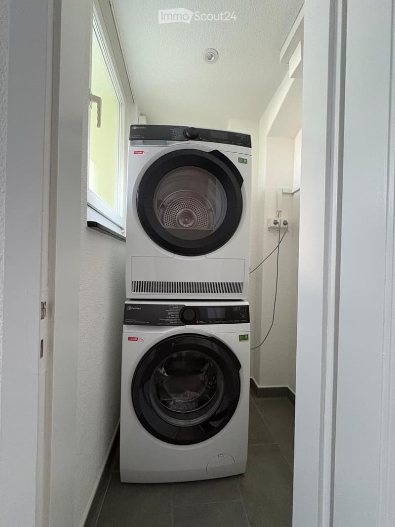 A small bathroom with a stacked washing machine and dryer unit, white walls, and a window on the left side.