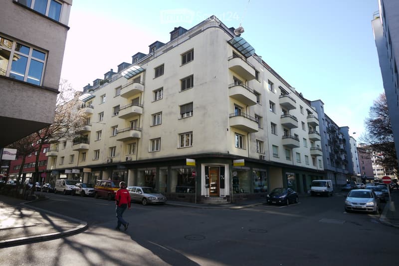 Multi-story residential building with balconies and storefronts at ground level, located on a street with parked cars and pedestrians.