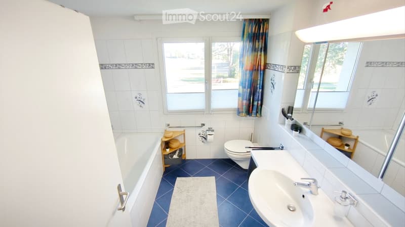 Bright bathroom with white tiles, blue floor tiles, bathtub, toilet, sink, mirror, and a small wooden shelf holding decorative items.