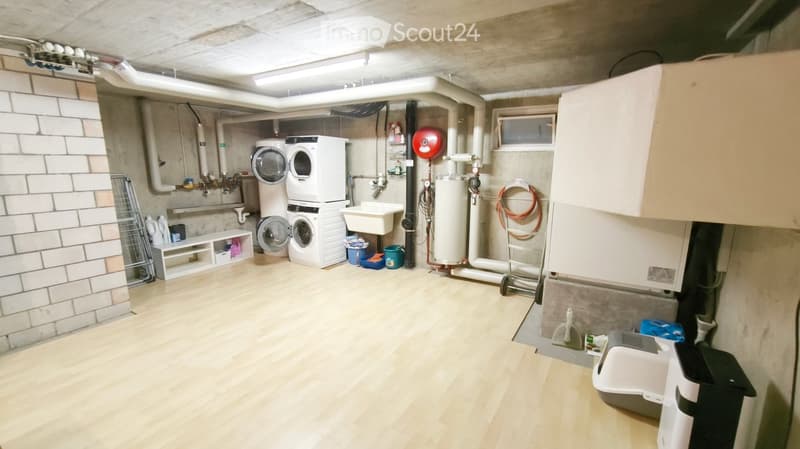 Well-lit basement utility room with wooden flooring, white brick wall, stacked washing machine and dryer, sink, water heater, storage shelves, and various utility items.