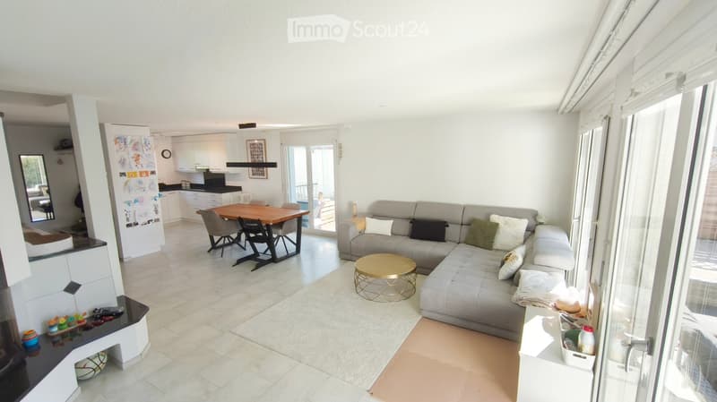 Modern living and dining area with a grey sectional sofa, coffee table, dining table with chairs, and a small kitchen area with cabinets and a refrigerator.