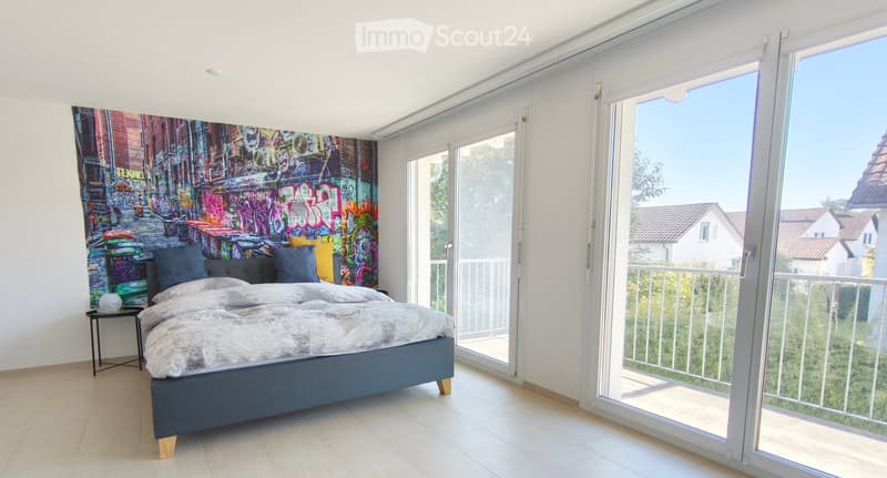 Modern bedroom with a large bed, nightstand, table lamp, and colorful graffiti-style wall art. Large sliding glass doors lead to a balcony with a view of the neighborhood.
