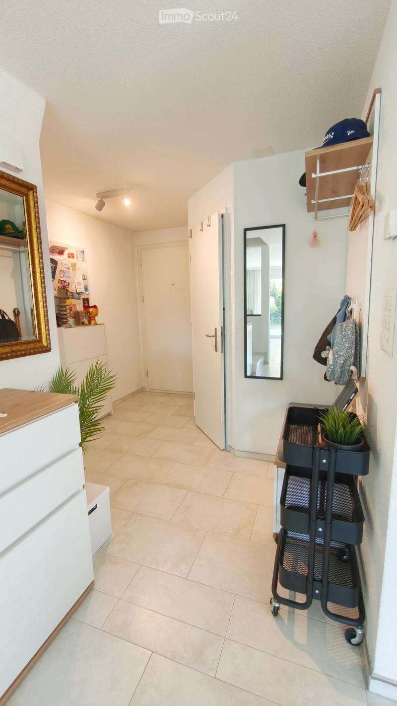 Bright hallway with tiled floor, white walls, and a coat rack with hanging items. A small cart with potted plants is positioned near the coat rack, and a mirror is mounted on the wall. A door leads to another room, and a small shelf with decorative items