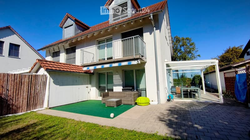 A modern two-story house with a balcony, surrounded by a well-maintained garden and patio area. The patio features a green artificial turf area with a wicker sofa and coffee table, a yellow inflatable pool, and a covered dining area with a table and chair