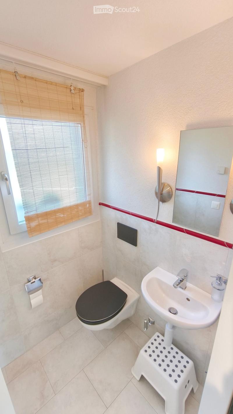 Modern bathroom with a toilet, sink, mirror, and a small white stool. Features a window with bamboo blinds and a wall-mounted light fixture.